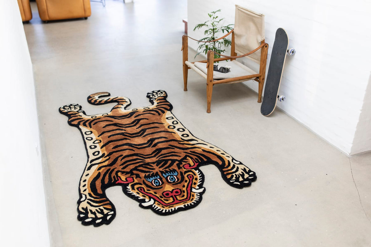 Tapis Tiger de Bongusta en forme de tigre posé sur un sol clair, dans une pièce blanche avec fauteuil en bois, plante et skateboard