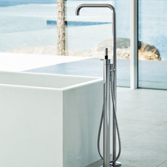 Mitigeur de baignoire au sol FS1 de VOLA en inox à côté d'une baignoire blanche, dans une salle de bains lumineuse ouverte sur la mer
